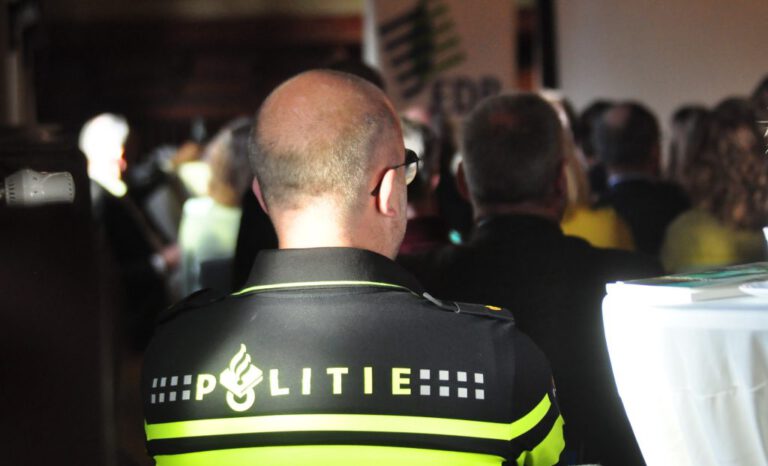 Im Hintergrund Publikum. Im Vordergrund Rückenansicht eines Mannes auf dessen Oberteil"Politie" steht.