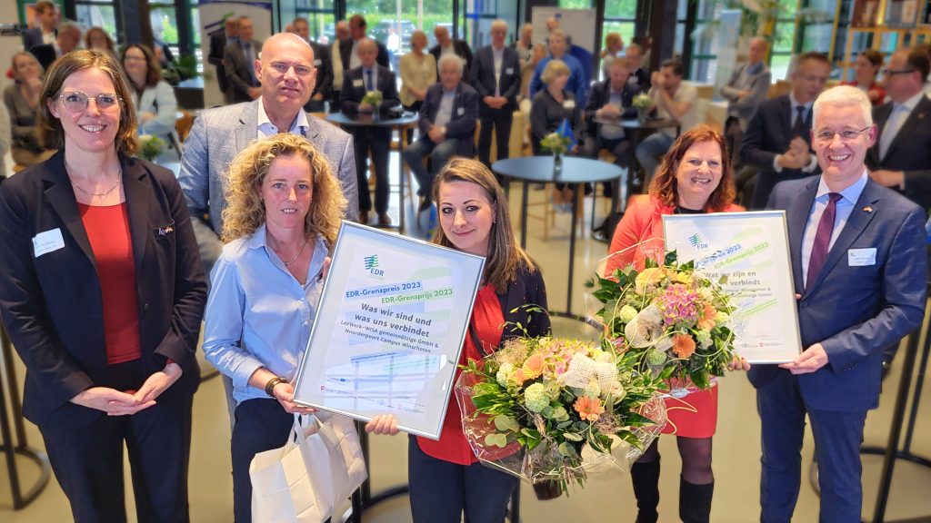Im Hintergrund Publikum. Im Vordergrund sechs Menschen, die zwei Blumensträuße und zwei Urkunden zeigen.