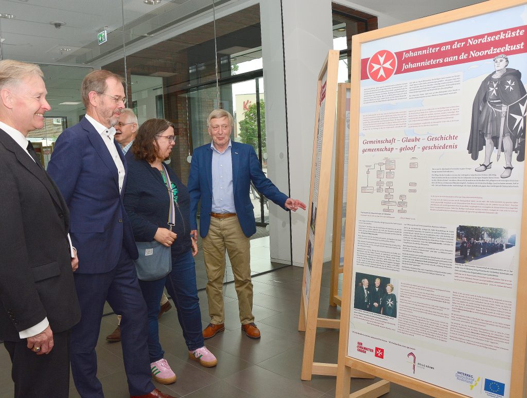 Fünf Menschen stehen vor einer Ausstellungstafel zum Thema "Johanniter an der Nordseeküste".