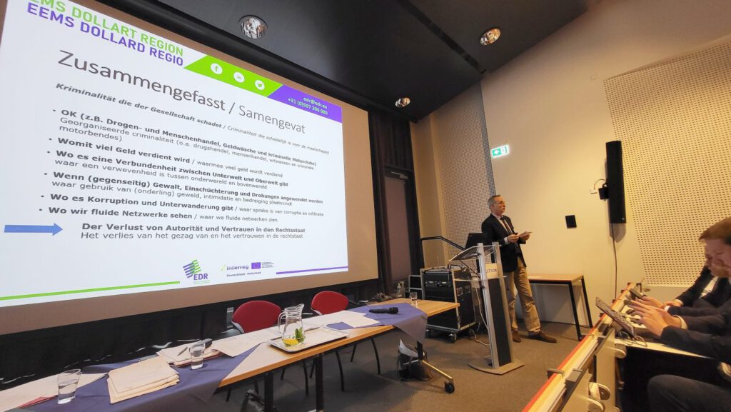 Ein stehender Mann schaut ins Publikum, hinter ihm eine Power-Point-Präsentation (Zusammenfassende Stichpunkte zu "Kriminalität, die der Gesellschaft schadet").