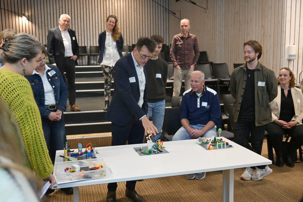 Das Bild zeigt eine Gruppe von Menschen, die an einer Präsentation teilnehmen. In einem modernen Raum mit Holzverkleidung stehen und sitzen sie um einen Tisch. Auf dem Tisch befinden sich zwei LEGO-Modelle, die von den Anwesenden gezeigt werden. Ein Mann, der ein formelles Outfit trägt, steht und erklärt das Modell, während er mit einer Hand darauf zeigt. Im Hintergrund ist eine kleine Gruppe von Personen zu sehen, die aufmerksam zuhören. Einige der Teilnehmer wirken interessiert, während andere lächeln oder in einer entspannten Haltung stehen.