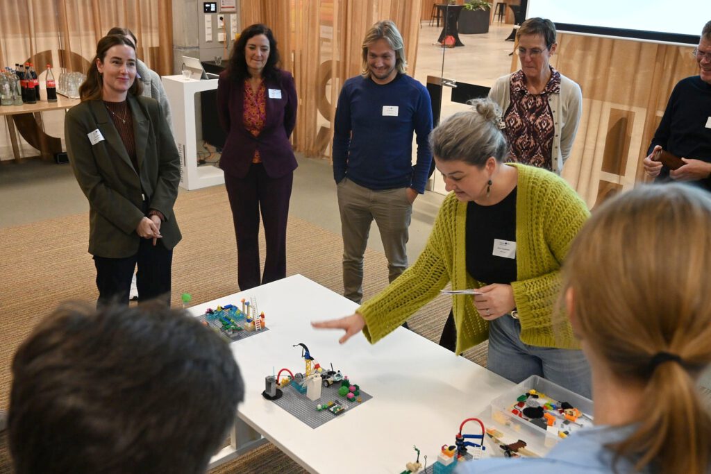 Menschen stehen um einen Tisch herum, auf dem Lego-Modelle stehen. Eine Frau hinter dem Tisch erklärt eins der Modelle.