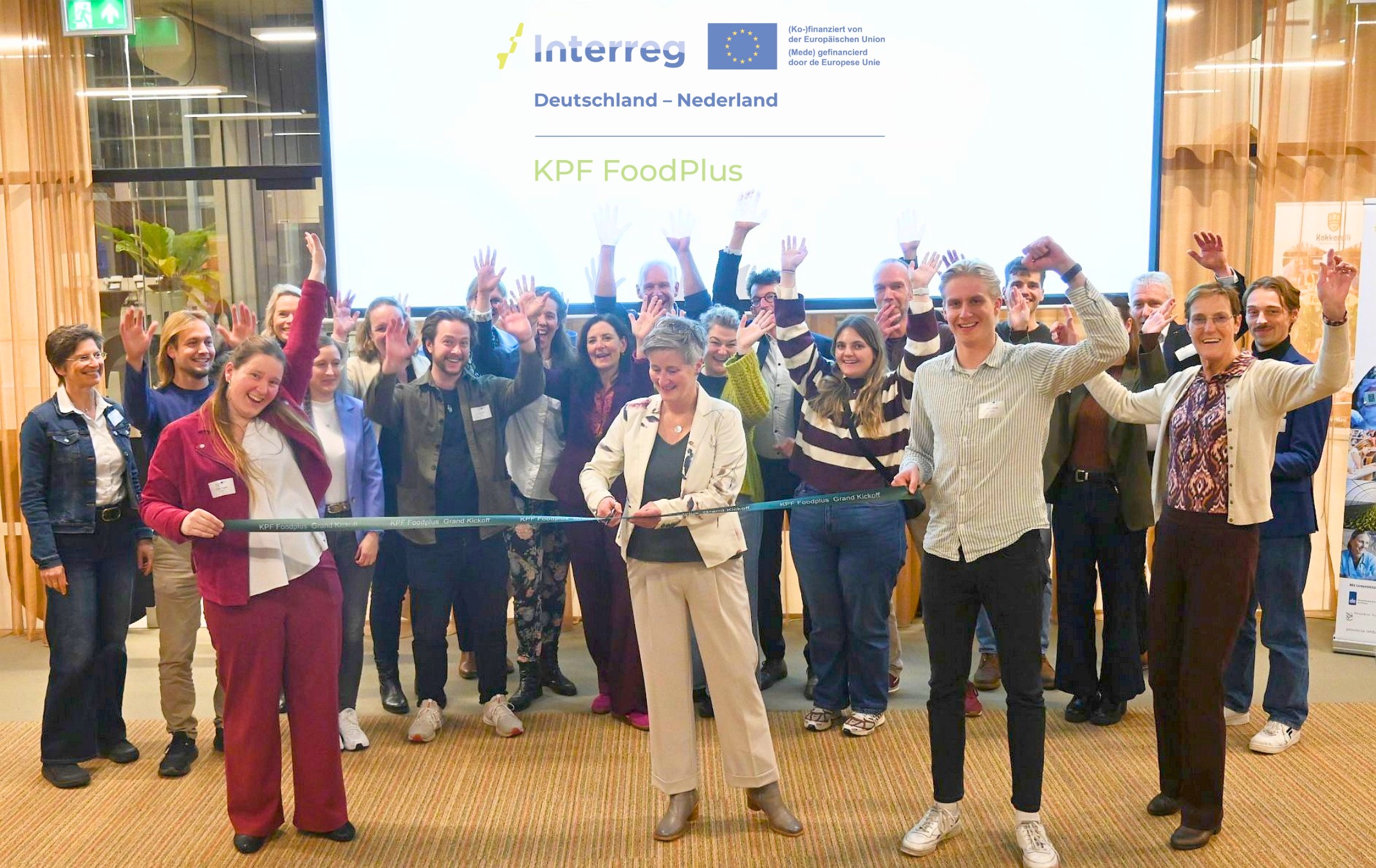 Menschen stehen vor einer Leinwand, im Vordergrund zerschneidet eine Frau ein Band, welches von einer Frau und einem Mann gespannt gehalten wird, als Zeichen für einen Start