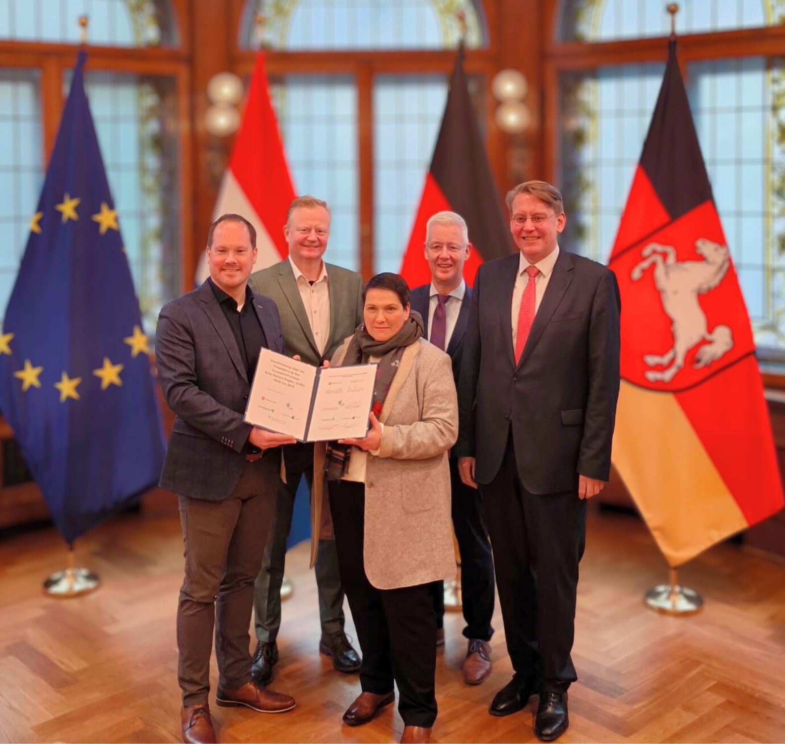 vier Männder und eine Frau, die Frau im Vordergrund hält zusammen mit einem Mann eine Zweiseitige Urkunde in den Händen und präsentiert diese. Alle sind in Business-Kleidung. Im Hintergrund sind die Flaggen von Niedersachsen, der EU, den Niederlanden und Deutschland. Vier mannen en één vrouw; de vrouw op de voorgrond houdt samen met een man een tweezijdige oorkonde vast en presenteert deze. Iedereen draagt zakelijke kleding. Op de achtergrond staan de vlaggen van Nedersaksen, de EU, Nederland en Duitsland.