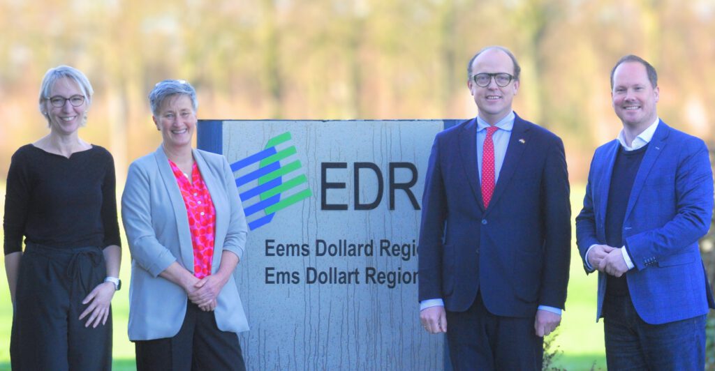 Fünf erwachsene Personen stehen im Freien vor einem Schild der EDR (Eems Dollard Regio) und blicken in die Kamera. Im Hintergrund ist eine unscharfe, herbstliche Landschaft zu sehen. Vijf volwassenen staan buiten voor een bord van de EDR (Eems Dollard Regio) en kijken in de camera. Op de achtergrond is een wazig herfstlandschap zichtbaar.