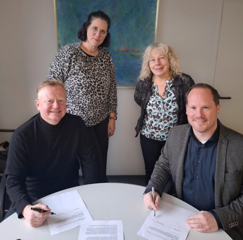 Im Hintergrund stehen zwei Frauen, vorne sitzen zwei Männer. Die Männer halten einen Stift in der Hand und haben Papiere vor sich auf einem Tisch liegen. Op de achtergrond staan twee vrouwen, vooraan zitten twee mannen. De mannen houden een pen in de hand en hebben papieren voor zich op een tafel liggen.