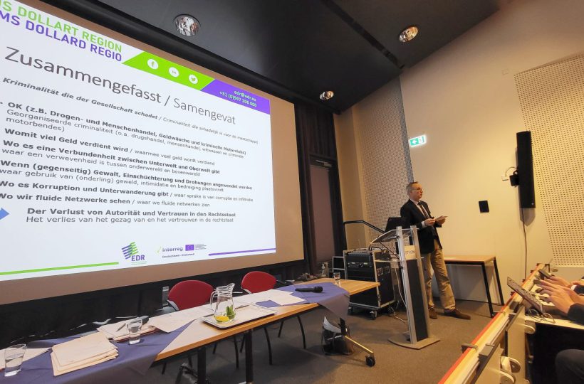 Ein stehender Mann schaut ins Publikum, hinter ihm eine Power-Point-Präsentation (Zusammenfassende Stichpunkte zu "Kriminalität, die der Gesellschaft schadet").