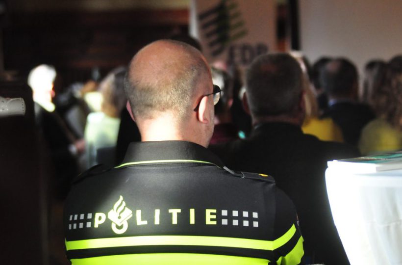 Im Hintergrund Publikum. Im Vordergrund Rückenansicht eines Mannes auf dessen Oberteil"Politie" steht.