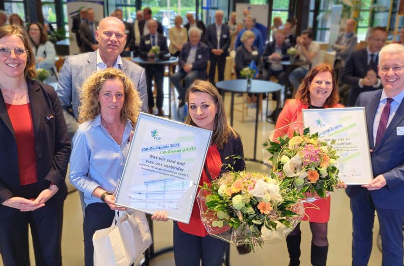 Im Hintergrund Publikum. Im Vordergrund sechs Menschen, die zwei Blumensträuße und zwei Urkunden zeigen.