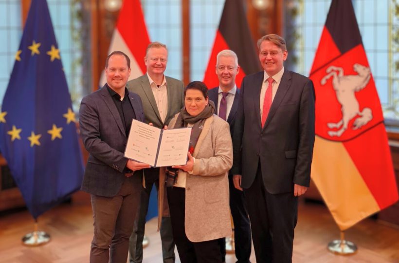 vier Männder und eine Frau, die Frau im Vordergrund hält zusammen mit einem Mann eine Zweiseitige Urkunde in den Händen und präsentiert diese. Alle sind in Business-Kleidung. Im Hintergrund sind die Flaggen von Niedersachsen, der EU, den Niederlanden und Deutschland. Vier mannen en één vrouw; de vrouw op de voorgrond houdt samen met een man een tweezijdige oorkonde vast en presenteert deze. Iedereen draagt zakelijke kleding. Op de achtergrond staan de vlaggen van Nedersaksen, de EU, Nederland en Duitsland.