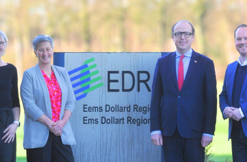 Fünf erwachsene Personen stehen im Freien vor einem Schild der EDR (Eems Dollard Regio) und blicken in die Kamera. Im Hintergrund ist eine unscharfe, herbstliche Landschaft zu sehen. Vijf volwassenen staan buiten voor een bord van de EDR (Eems Dollard Regio) en kijken in de camera. Op de achtergrond is een wazig herfstlandschap zichtbaar.