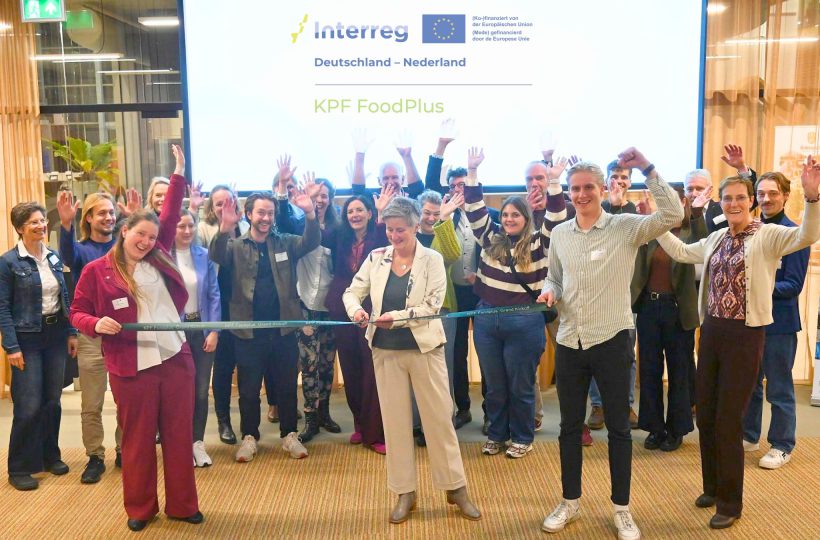 Menschen stehen vor einer Leinwand, im Vordergrund zerschneidet eine Frau ein Band, welches von einer Frau und einem Mann gespannt gehalten wird, als Zeichen für einen Start