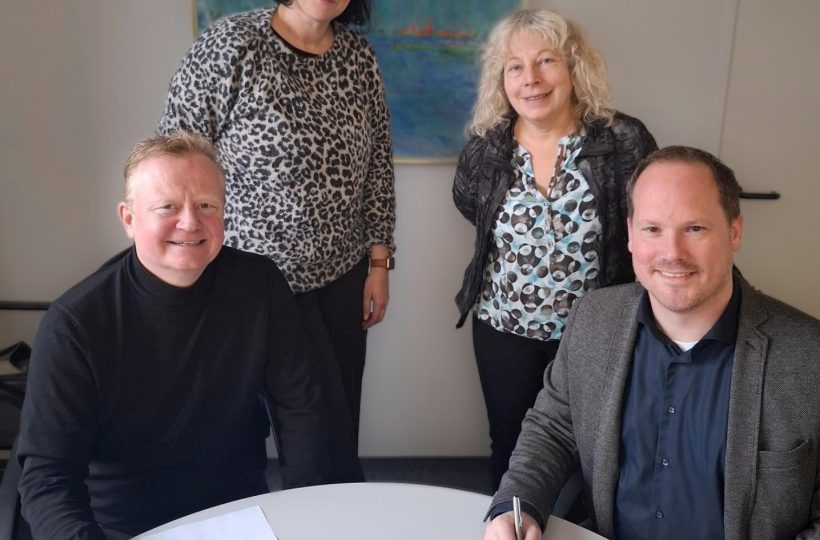 Im Hintergrund stehen zwei Frauen, vorne sitzen zwei Männer. Die Männer halten einen Stift in der Hand und haben Papiere vor sich auf einem Tisch liegen. Op de achtergrond staan twee vrouwen, vooraan zitten twee mannen. De mannen houden een pen in de hand en hebben papieren voor zich op een tafel liggen.