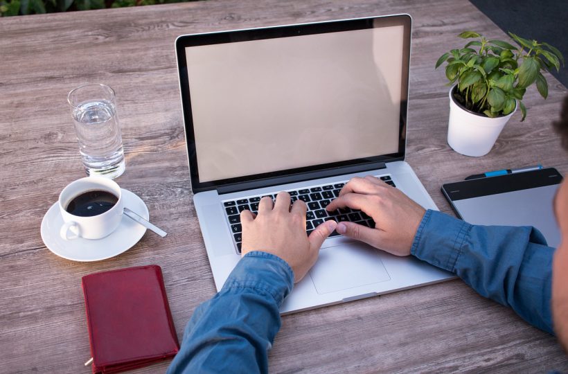 Person tippt auf Laptop, daneben Kaffee, Wasser, Notizbuch und Pflanzenkübel.